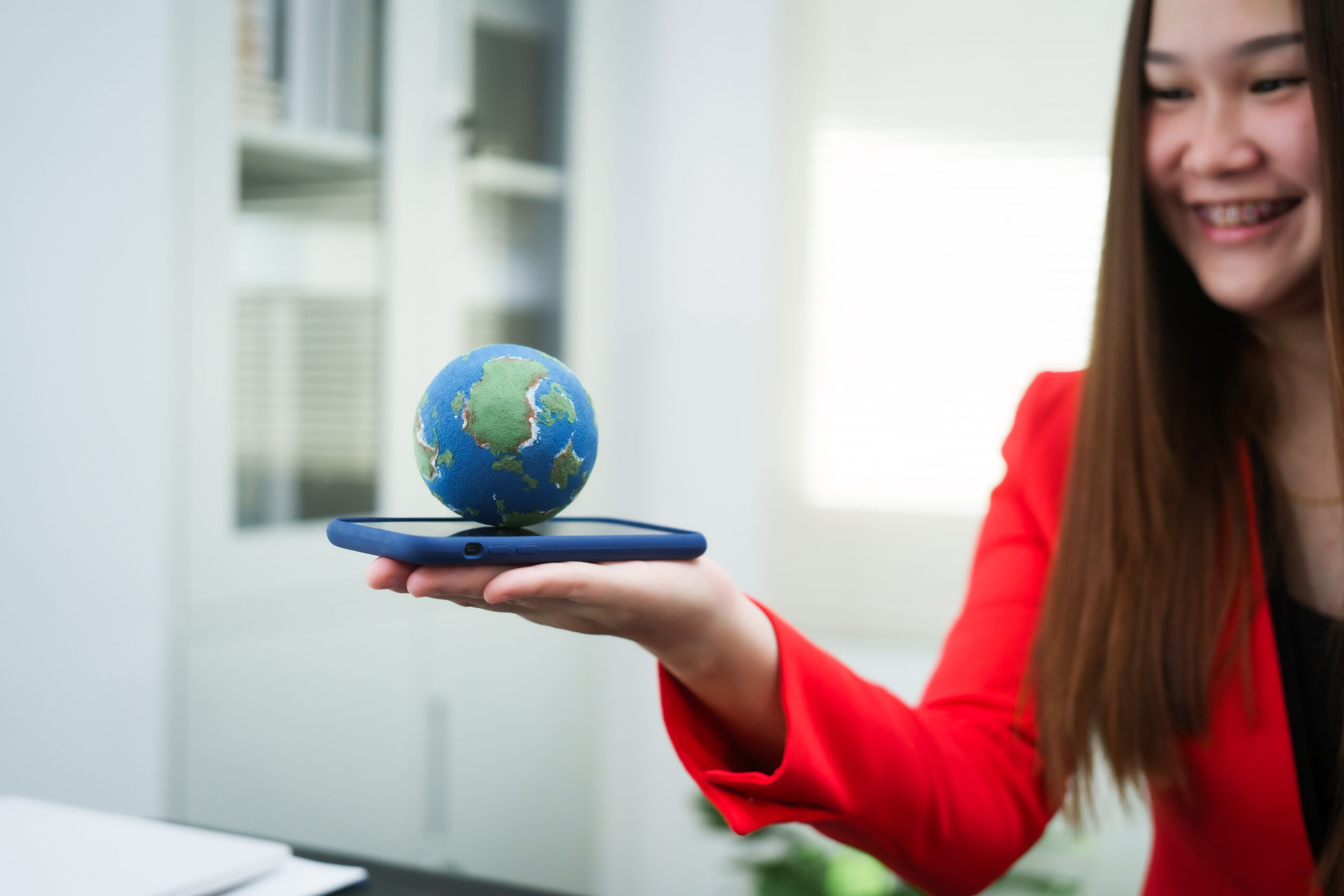 A smiling young professional in a red blazer holds a smartphone with a small globe on top, symbolising global opportunities and UK visa options.
